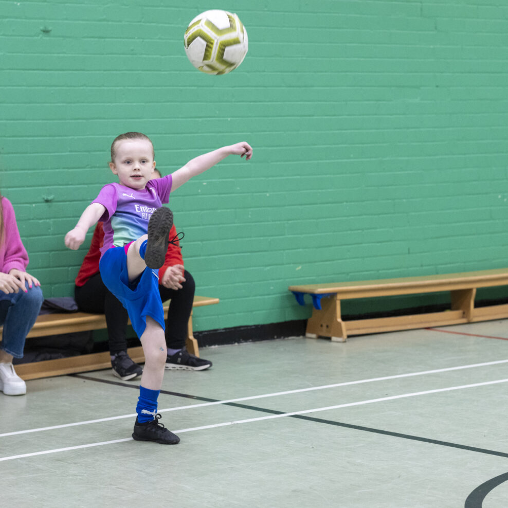 A young member at The Bronte Youth and Community Centre playing football in our sports hall as other young people and staff spectate. Based in Liverpool City Centre.